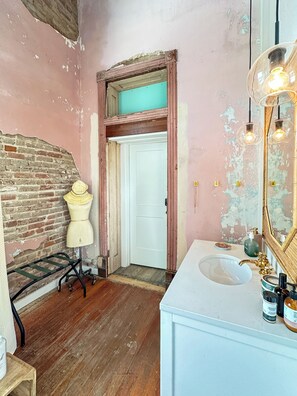 A bathroom with a rustic brick wall, a mannequin, a white door, a wooden floor, and a vintage-style mirror with lights. - 3E Sunrise View (Honey Grove)