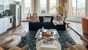 A modern kitchen with stainless steel appliances, a dark blue sofa with cushions, a glass coffee table with books and a bowl, and a view of the outdoors through large windows. - 3E Sunrise View (Honey Grove)