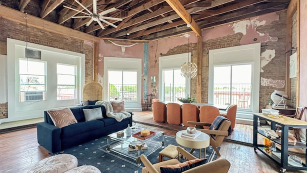 A spacious living room with a high ceiling, exposed beams, and large windows. There is a blue sofa, a round coffee table, and several chairs. - 3E Sunrise View (Honey Grove)