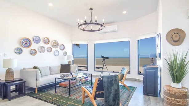 Ein geräumiges Wohnzimmer mit großem Fenster mit Blick auf einen Strand, ein bequemes Sofa, ein Couchtisch mit Büchern und dekorative Teller an der Wand.