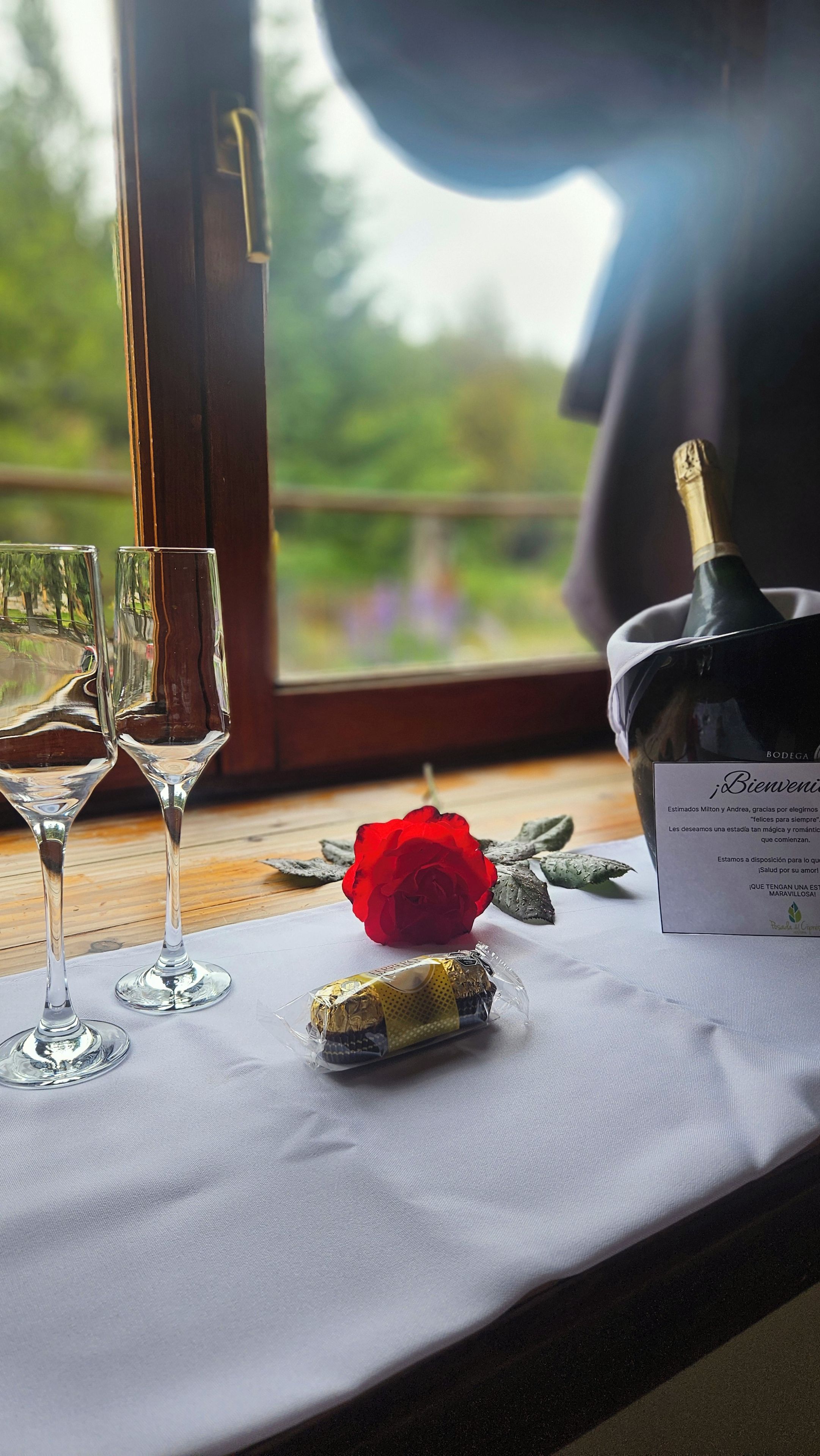 A table with two glasses, a bottle of champagne, a wrapped chocolate bar, and a red rose on a white tablecloth.