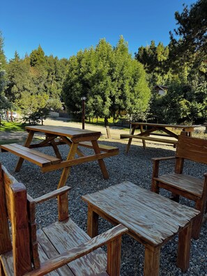 A picnic area with wooden benches and picnic tables, surrounded by gravel and trees. - POSADA DEL CIPRES (Villa La Angostura)