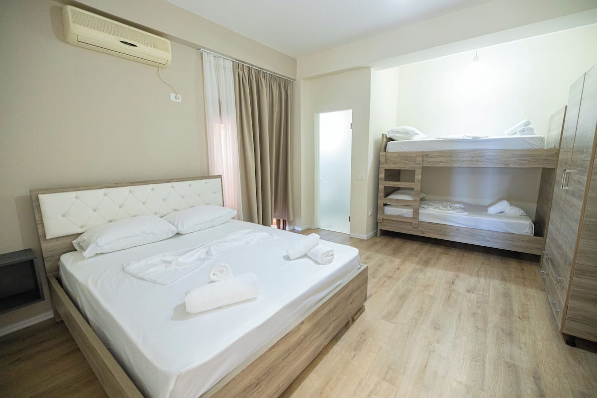 A hotel room with a large bed, a smaller bunk bed, wooden flooring, a built-in wardrobe, and a ceiling-mounted air conditioner.