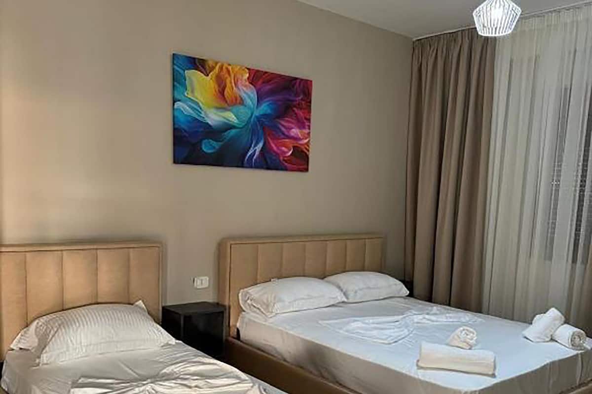 A hotel room with two beds, a bedside table, a lamp, and a colorful abstract painting on the wall.