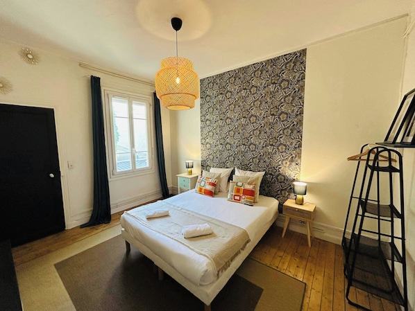 A bedroom with a bed, a hanging light fixture, a bedside table with a lamp, a wardrobe, and a window with curtains.