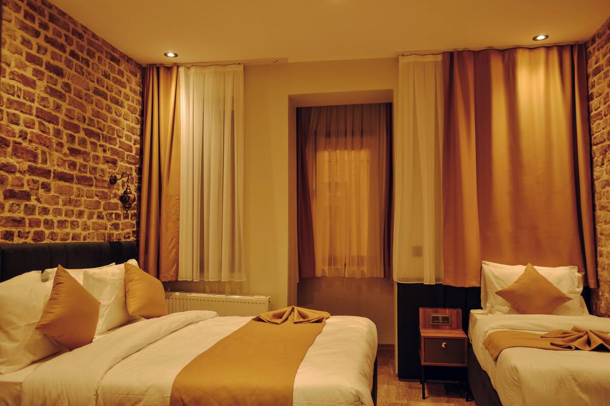 Une chambre d’hôtel avec un mur en briques, deux lits avec une literie blanche et des touches de couleur marron, une table de chevet en bois et une fenêtre avec des rideaux.