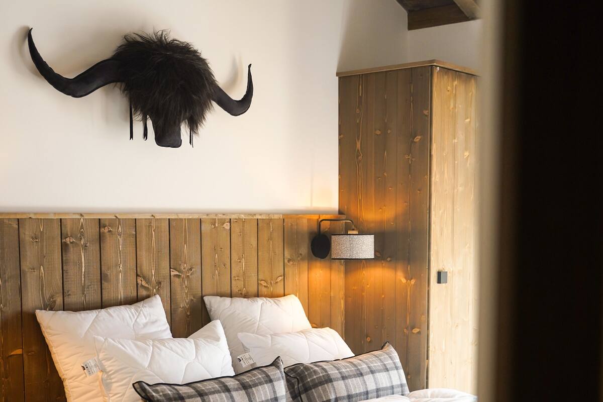 Ein Schlafzimmer mit einem hölzernen Kopfteil, einem Bett mit weißer und karierter Bettwäsche sowie einem aufgehängten Tierkopf an der Wand.