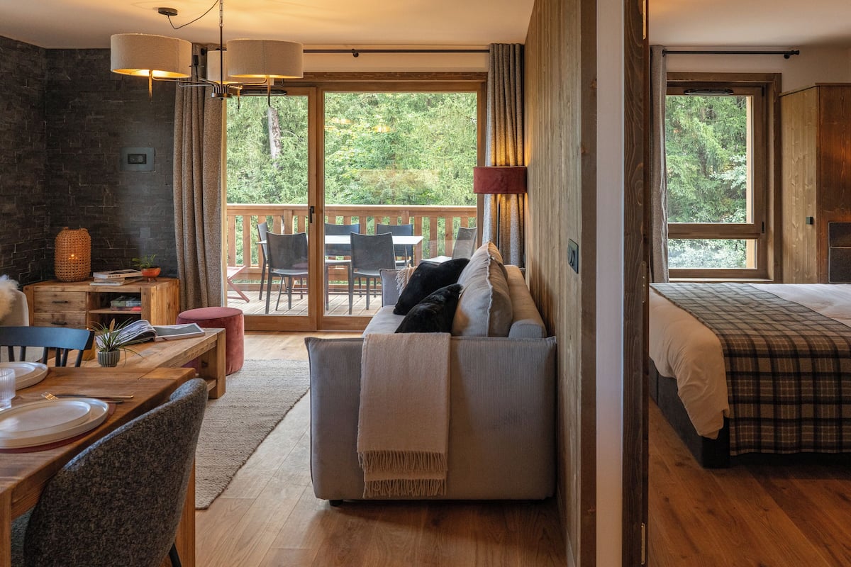Ein gemütliches Zimmer mit Holztisch, einer Couch, einem Bett mit kariertem Überwurf und Blick ins Freie durch große Fenster.