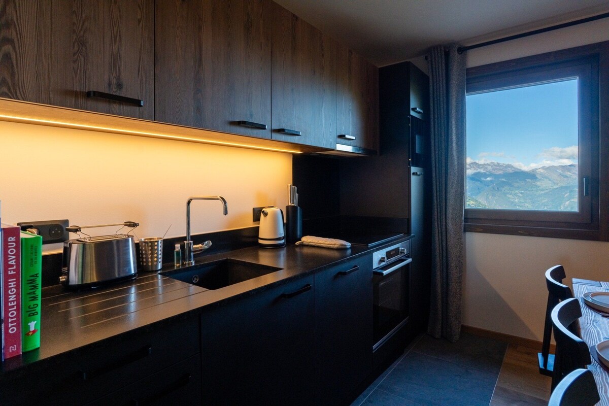 Eine moderne Küche mit dunklen Schränken, einer Edelstahl-Dunstabzugshaube und einem Fenster mit Blick auf die Berge.