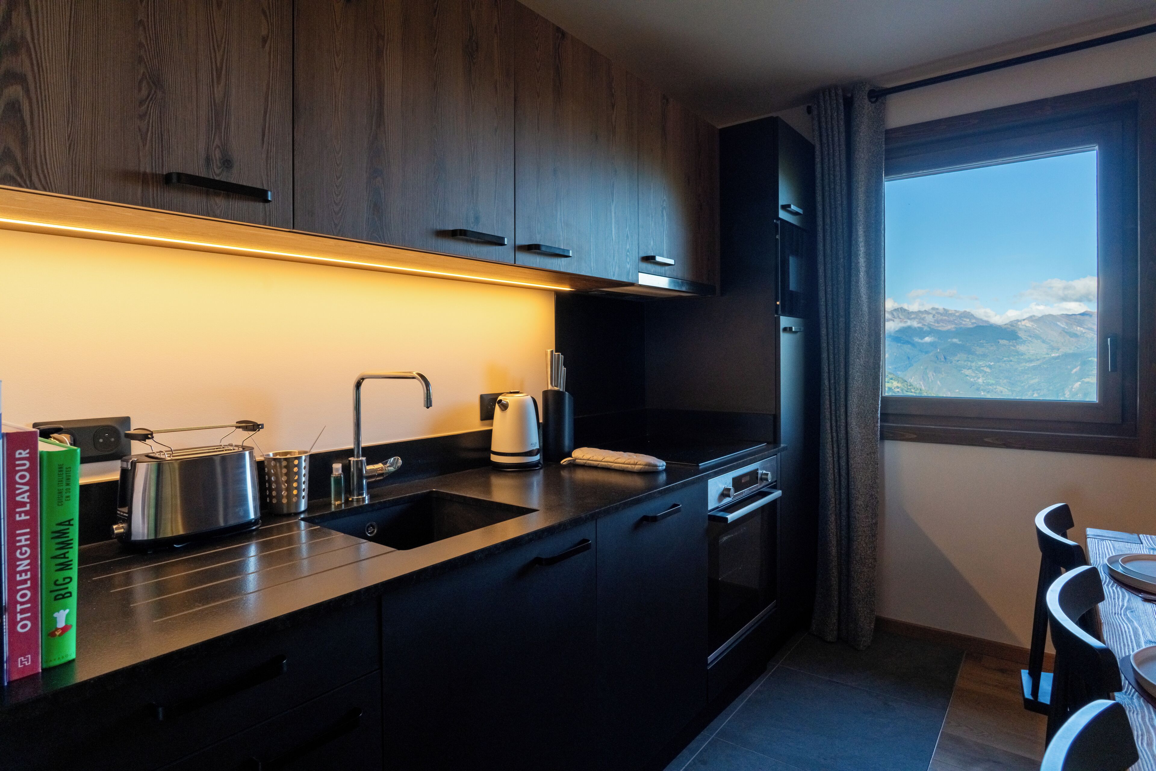 A modern kitchen with dark cabinetry, a stainless steel toaster, a kettle, and a window offering a mountain view.