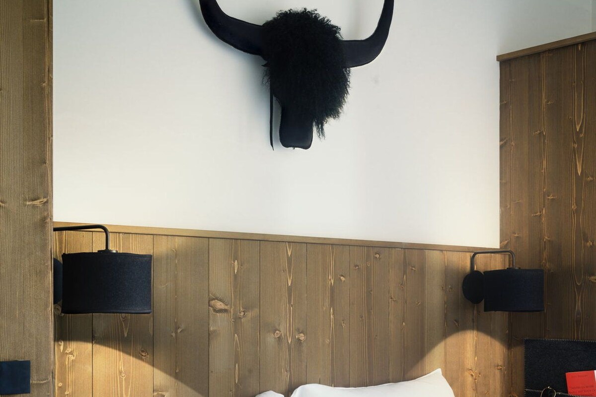 Ein Schlafzimmer mit einem hölzernen Kopfteil, zwei Nachttischlampen, einem Bett mit weißer und grauer Bettwäsche und einem aufgehängten Stierkopf an der Wand.