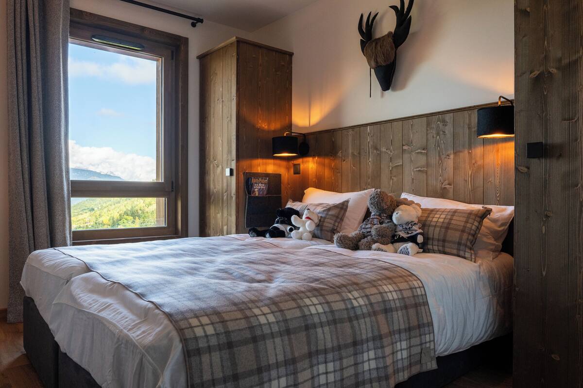 Ein Schlafzimmer mit einem großen Bett, einem Holzkopfteil, einem aufgehängten Hirschgeweih, einem Fenster mit Blick und einem Teddybären auf dem Bett.