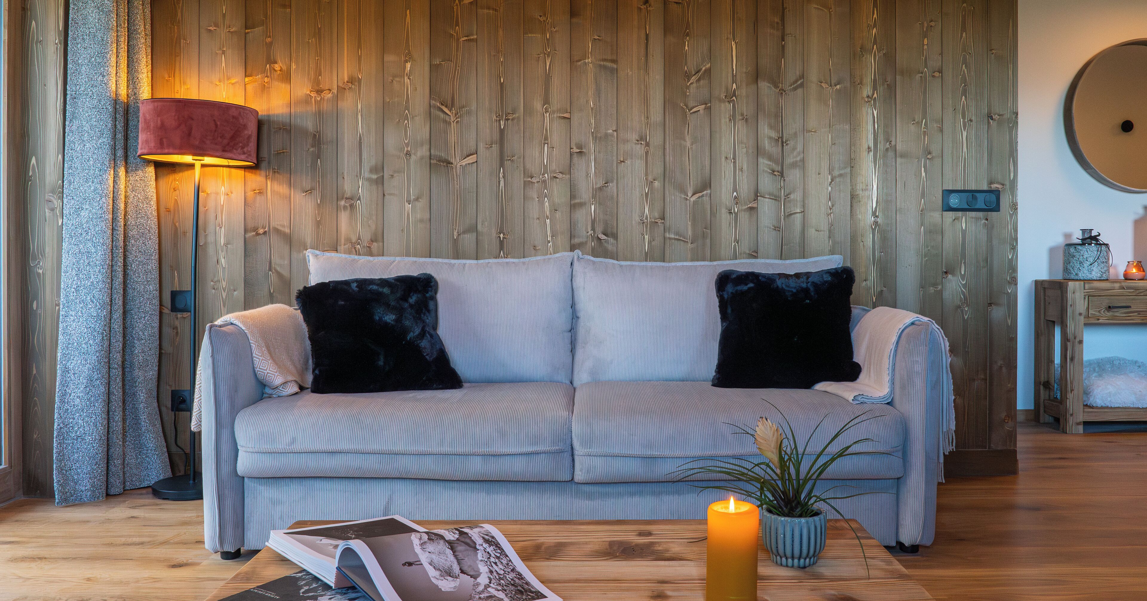 A living room with a light blue sofa, two black cushions, a wooden wall, a floor lamp, a table with a candle, and a small plant.