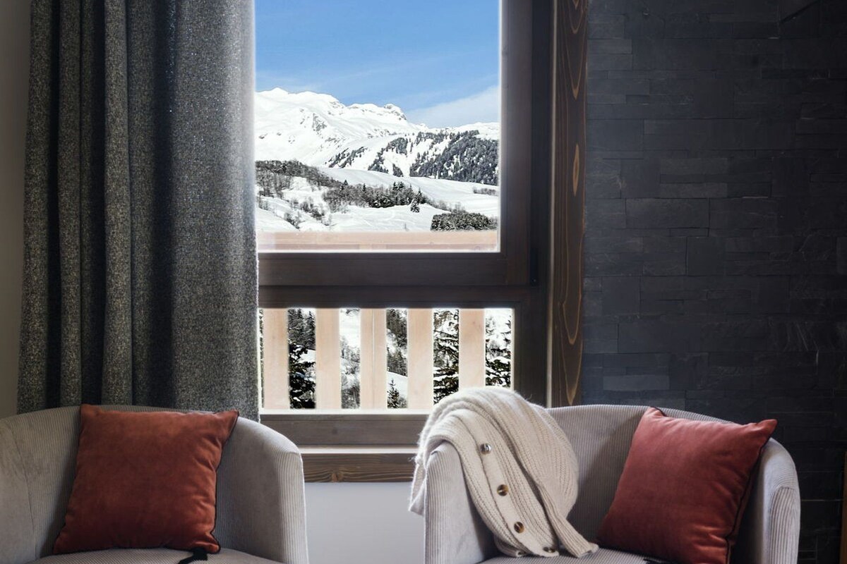 Zwei graue Sessel mit roten Kissen, ein Holztisch mit Orangensaft, Gläsern und einem Buch, und ein Fenster mit Blick auf einen verschneiten Berg.