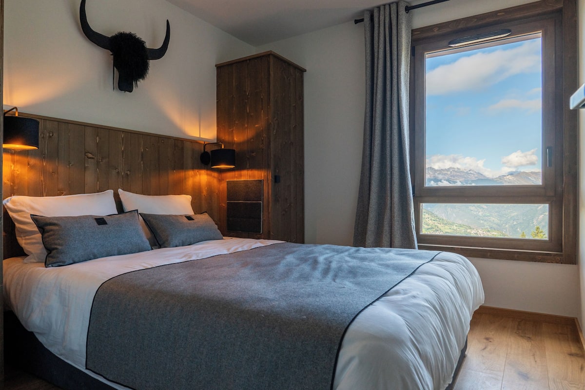 Ein Schlafzimmer mit Bett, Holz-Kopfteil, Wandlampen, einem großen Fenster mit Bergblick und einem Holzschrank.