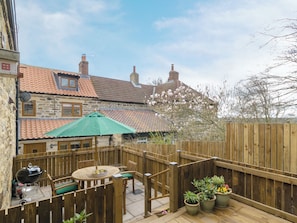 A wooden deck with a table and chairs, surrounded by a wooden fence, and a stone building with a tiled roof in the background.