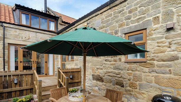 A stone building with a wooden deck, a round wooden table with chairs, a green umbrella, and a barbecue grill. - 2 bedroom accommodation in Grinkle, near Staithes (Grinkle, near Staithes)