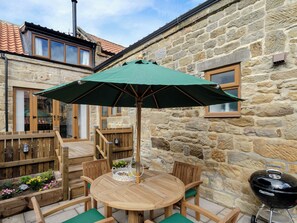 A stone building with a wooden deck, a round wooden table with chairs, a green umbrella, and a barbecue grill.