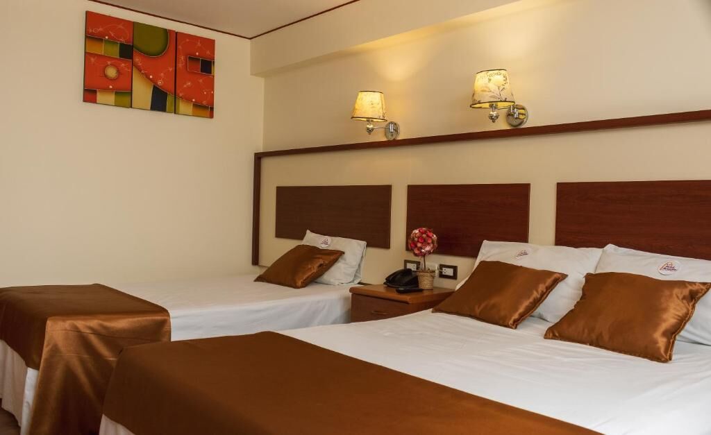 A hotel room with two beds, each with brown headboards and white bedding. There are two bedside tables with lamps, a wall-mounted artwork, and a small vase with flowers.