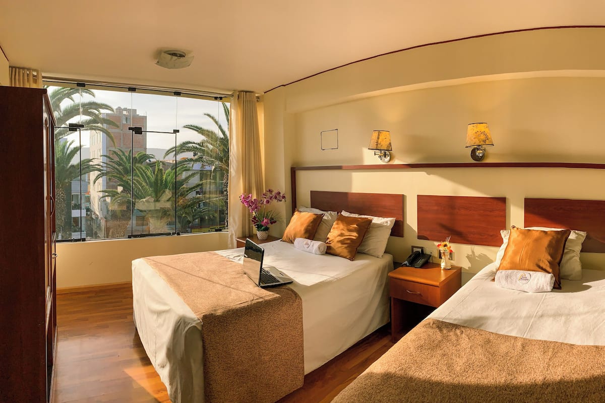 A hotel room with two beds, a desk, a laptop, a nightstand with a lamp, and a view of palm trees and buildings.