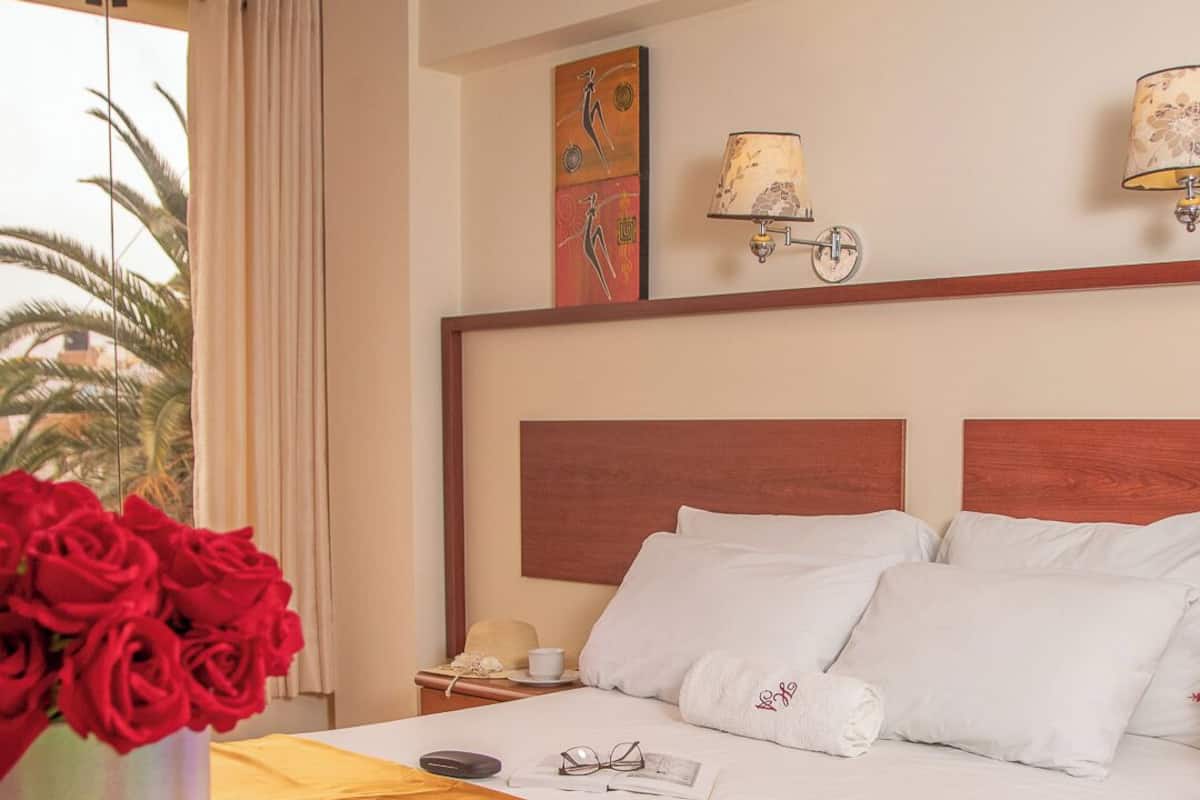 A hotel room with a bed, a bedside table with a lamp, a vase of red roses, and a window with curtains.