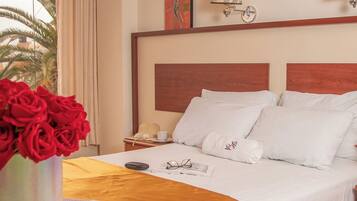 A hotel room with a bed, a bedside table with a lamp, a vase of red roses, and a window with curtains.