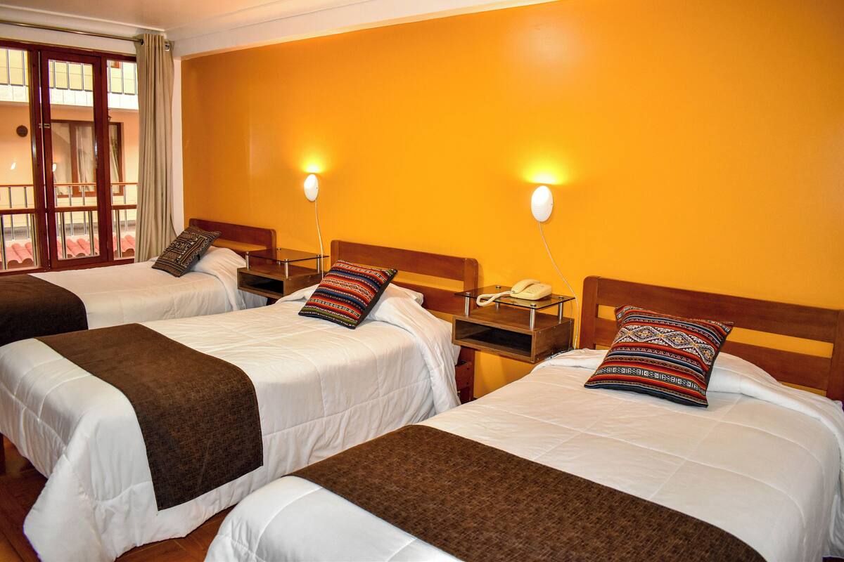 Habitación de hotel con dos camas, cada una con una manta marrón y almohadas estampadas, ropa de cama blanca y mesitas de noche con lámparas y un teléfono.