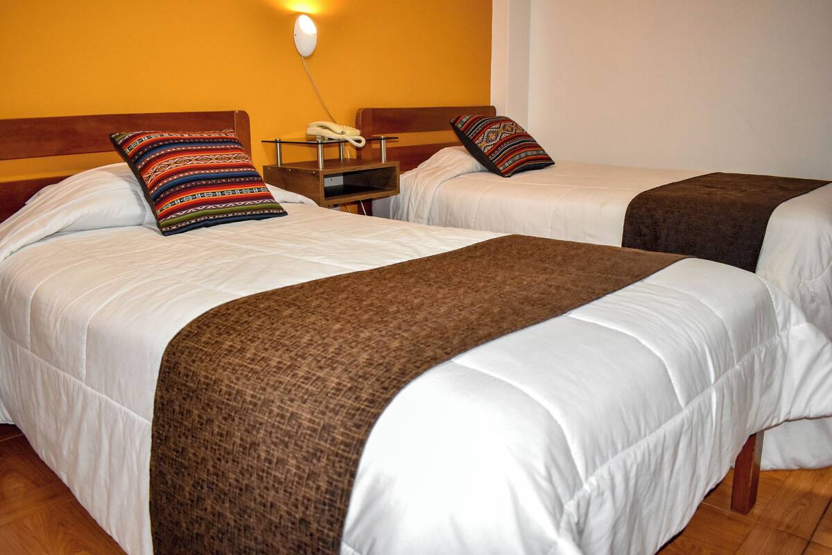Dos camas individuales con ropa blanca y mantas marrones, una mesita de noche con teléfono, y una lámpara de pared.