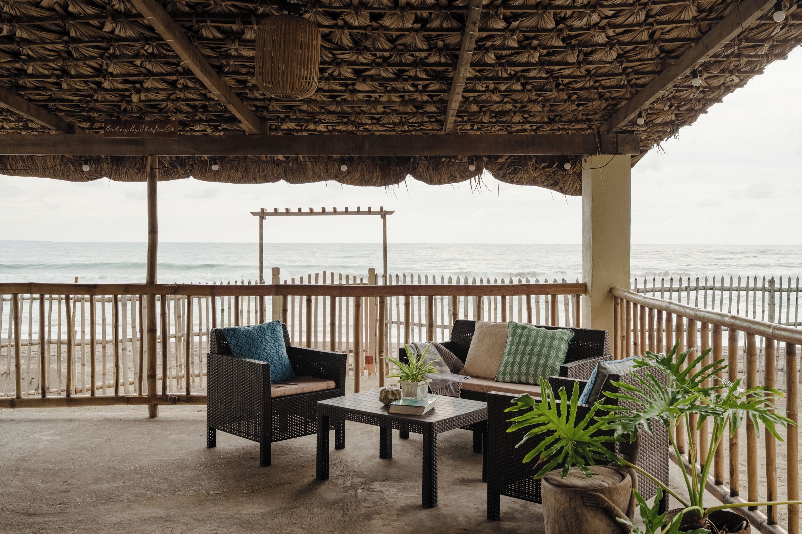 A covered outdoor seating area with a view of the sea, featuring wicker furniture and potted plants.