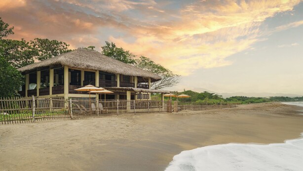 A beachfront property with a thatched-roof building, outdoor seating, and a view of the ocean. - Serene beach front Villa near Vigan (San Juan)