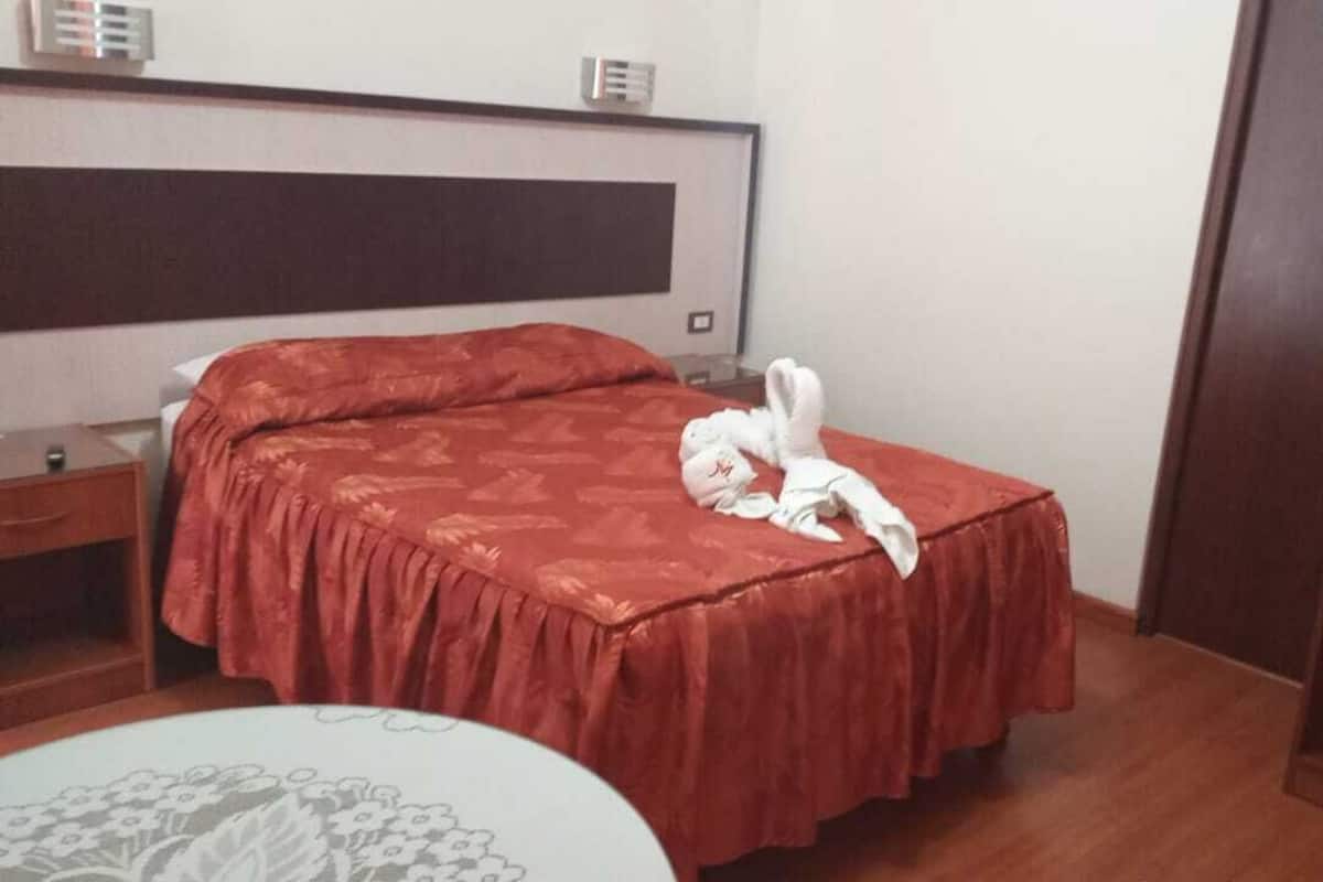A hotel room with a bed, a bedside table, a nightstand, a chair, and a small table with a floral tablecloth.