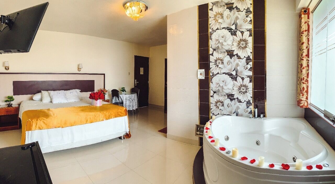A hotel room with a bed, a sitting area with a table and chairs, a large bathtub with rose petals, and a wall with a floral pattern.