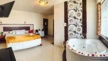 Habitación de hotel con cama, zona de estar con mesa y sillas, una bañera grande con pétalos de rosa y una pared con estampado floral.