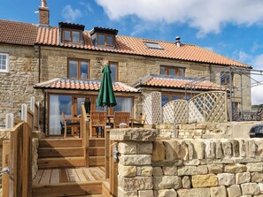 Un bâtiment en pierre avec un toit en tuiles, des marches en bois et une terrasse en bois équipée de chaises.