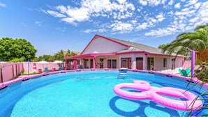 Una piscina con baldosas rosadas y azules, una casa de techo rosa y un cielo azul despejado.