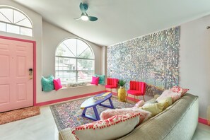A cozy living room with a pink door, a large window, a ceiling fan, and a wall with a mosaic pattern.