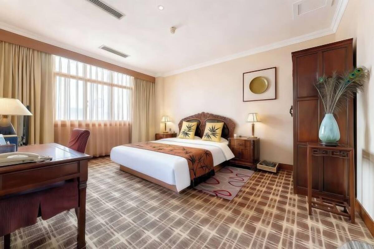 Hotelkamer met een bed, nachtkastjes, een bureau, een stoel, een groot raam met gordijnen, een wandklok en een decoratieve vaas.