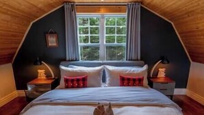 A bedroom with a sloped ceiling, a large window, a bed with pillows, and bedside lamps.