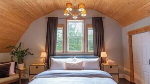 Un dormitorio con techo inclinado, una cama con sábanas blancas, dos mesitas de noche con lámparas y un ventanal con cortinas.