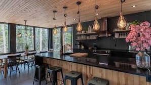 Une cuisine moderne avec un plafond en bois, des luminaires suspendus, un grand îlot central avec des tabourets et une vue sur la verdure grâce à de grandes fenêtres.