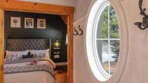 Ein Schlafzimmer mit einem gepolsterten Kopfteil, einem Bett mit vielen Kissen, einem Nachttisch mit Lampe und einem großen Fenster mit Blick auf Bäume.