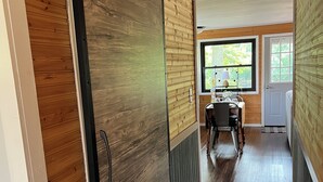 A hallway with a sliding door, a dining area with a table and chairs, and a window with a view of greenery. -  Little River House on Henry, on the serene banks of  the Iroquois River (Ashkum)