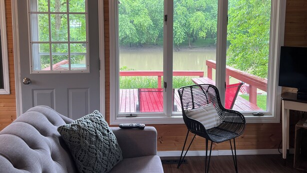 A room with a wooden interior, a grey sofa with a patterned pillow, a black chair with a striped pillow, and a large window overlooking a deck and greenery. -  Little River House on Henry, on the serene banks of  the Iroquois River (Ashkum)