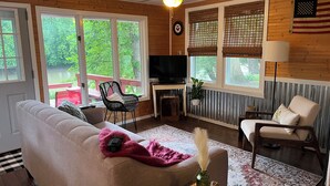 A cozy living room with a sofa, a chair, a TV, and a view of a deck and greenery. -  Little River House on Henry, on the serene banks of  the Iroquois River (Ashkum)
