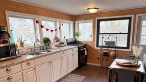 A modern kitchen with white cabinets, a granite countertop, and a black stove. There are windows with curtains, a sink, and a dining area with a chair and table. -  Little River House on Henry, on the serene banks of  the Iroquois River (Ashkum)
