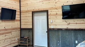 Habitación con paredes de madera, una puerta blanca, televisor montado, zapatero y sofá.