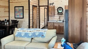 Una sala de estar acogedora con un sofá, una mesa de centro y un televisor. Se observa un techo de madera y una cocina al fondo.