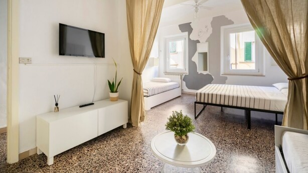 Ein Zimmer mit einem weißen Bett, einer Couch, einem an der Wand montierten Fernseher und einem kleinen Tisch mit einer Pflanze.