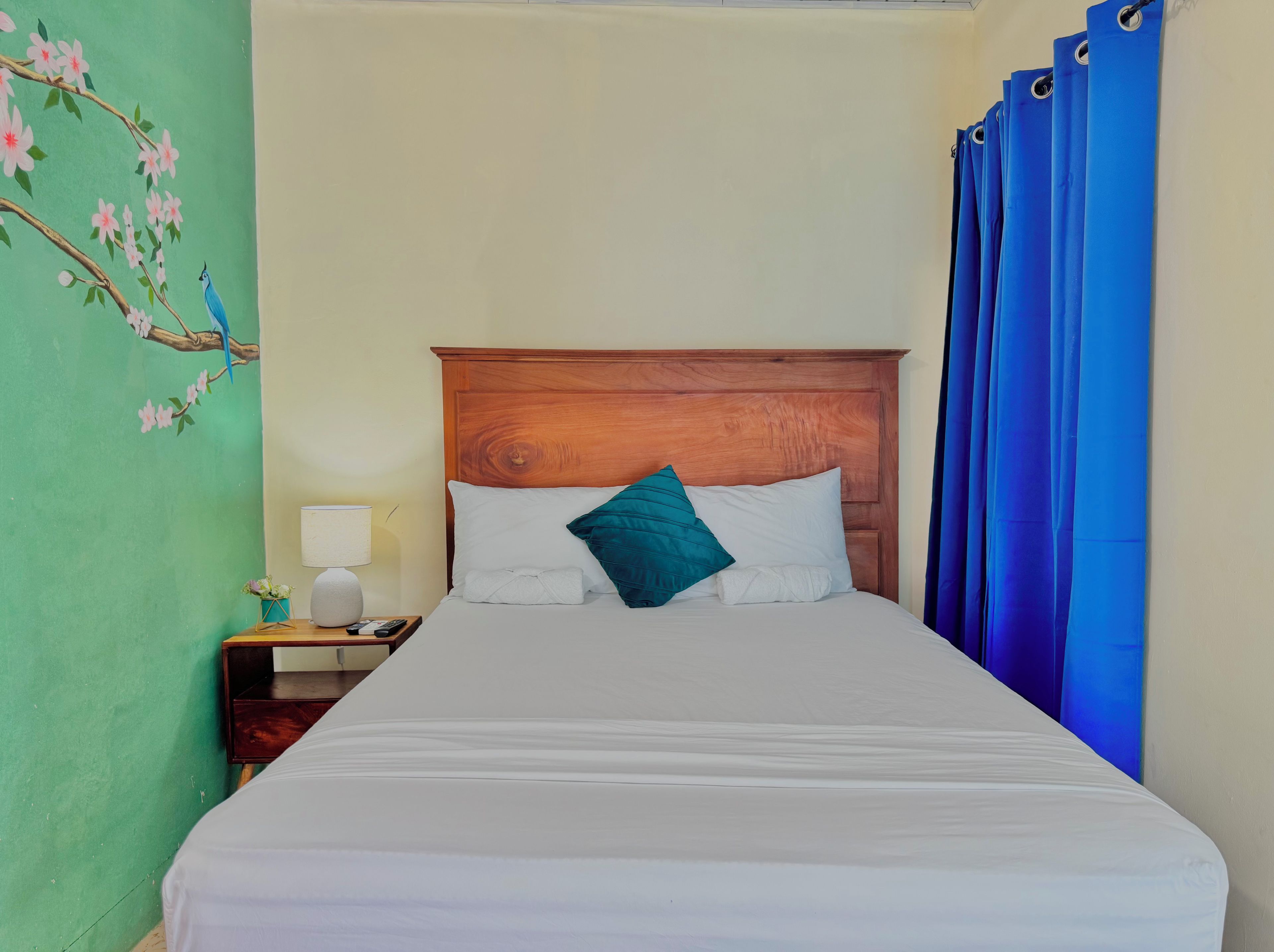 A hotel room with a wooden headboard, a bed with white linens, a teal pillow, a nightstand with a lamp, and a wall with a floral mural.