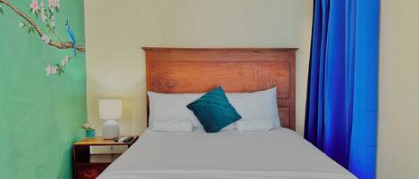 A hotel room with a wooden headboard, a bed with white linens, a teal pillow, a nightstand with a lamp, and a wall with a floral mural.